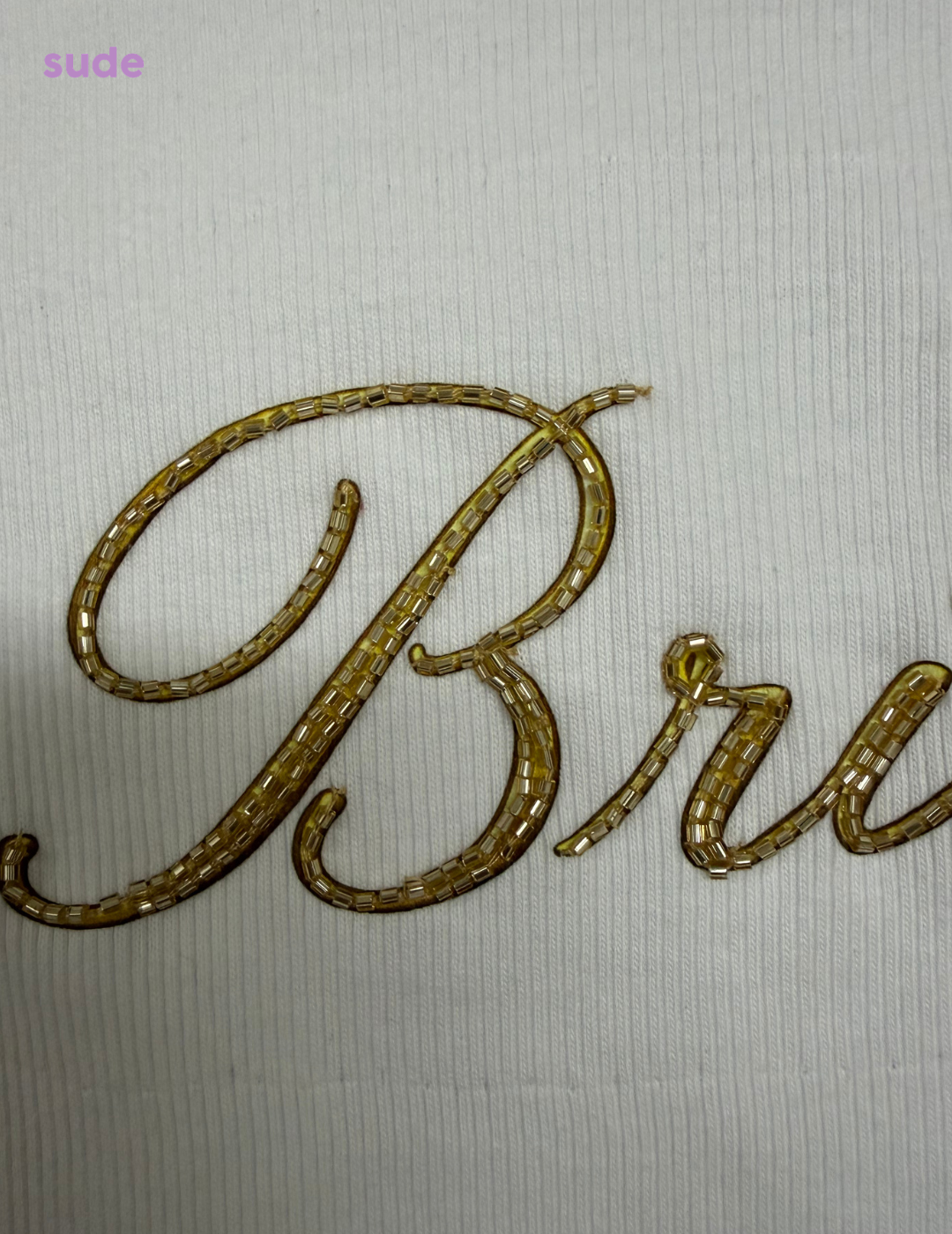 A close-up of a gold metallic embroidered script lettering on a textured off-white fabric, with the word 'Brun' partially visible.