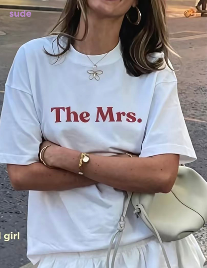 A woman wearing a white oversized T-shirt with the words 'The Mrs.' printed in red across the chest, styled with gold jewelry and a light-colored handbag.